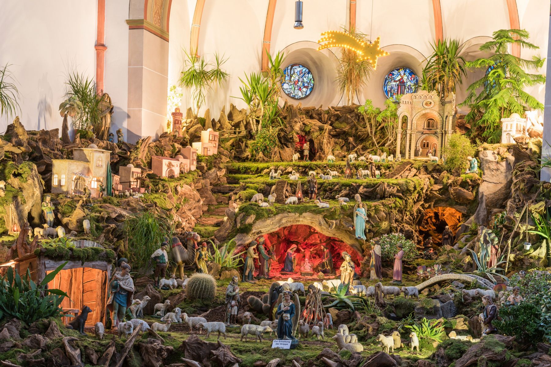 Eine detailreiche und weitläufige Krippe in einer Kirche mit vielen Figuren von Menschen, Tieren, Gebäuden und Grünpflanzen, mit Buntglasfenstern im Hintergrund und einem hellen Stern darüber.